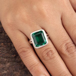 May include: A silver ring featuring a large, emerald-cut, deep green gemstone. The gemstone is set in a silver band with a square, slightly rounded bezel. The ring is shown on a person's finger.