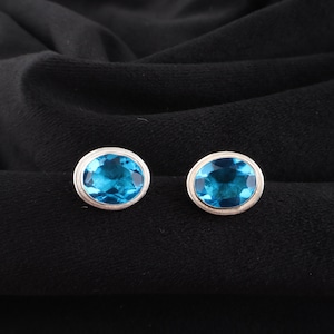 May include: A pair of oval-shaped, blue gemstone stud earrings with a silver-colored bezel setting. The earrings are displayed on a black fabric background, highlighting the vibrant blue color of the gemstones.