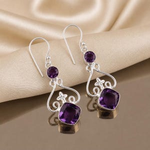 May include: A pair of silver drop earrings featuring a round purple amethyst stone at the top and a larger cushion-cut purple amethyst stone at the bottom, connected by a decorative silver wire frame with a small four-petal flower accent.