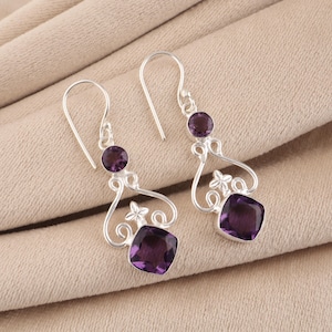 May include: A pair of silver dangle earrings with purple amethyst gemstones. Each earring features a hook, a small round stone, a decorative silver filigree design, and a larger square-cut amethyst. The earrings are displayed on a neutral fabric.