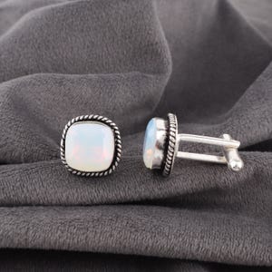 May include: A pair of square cufflinks with a white opal stone set in a silver-toned metal frame. The frame has a rope-like border. The cufflinks are displayed on a gray fabric surface.