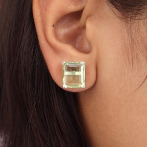 May include: A close-up of a light green rectangular gemstone stud earring. The gemstone is set in a silver metal setting and is worn on the ear. The earring has a rectangular shape with a light green color.