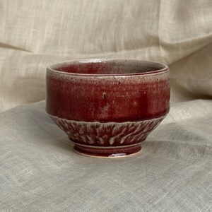 Op de afbeelding: Handgemaakte keramische kom met een dieprode glazuur. De kom heeft een gestructureerd, geribbeld ontwerp op de onderste helft en een kleine, cirkelvormige basis. De rand van de kom heeft een lichtere, gespikkelde kleur. Ideaal voor het serveren van soepen, ontbijtgranen of snacks.