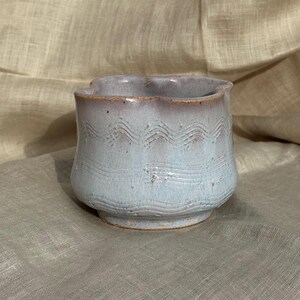 Op de afbeelding: Een handgemaakte keramische beker met een lichtblauwe glazuur en een golvend patroon in het midden. De rand heeft een geschulpte rand en een roodbruine tint. De beker staat op een kleine, afgeronde basis.