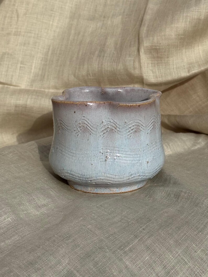 Op de afbeelding: Een handgemaakte keramische beker met een lichtblauwe glazuur en een golvend patroon in het midden. De rand heeft een geschulpte rand en een roodbruine tint. De beker staat op een kleine, afgeronde basis.