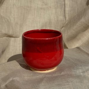 Op de afbeelding: Een handgemaakte, rode keramische beker met een afgeronde vorm. De beker heeft een glanzende afwerking en een lichtere basis. De binnenkant en het bovenste deel van de beker zijn een diepere tint rood, waardoor een gradi&euml;nteffect ontstaat.
