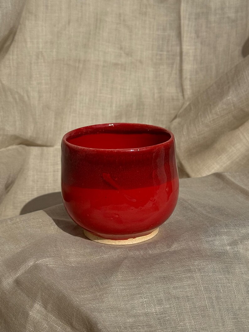 Op de afbeelding: Een handgemaakte, rode keramische beker met een afgeronde vorm. De beker heeft een glanzende afwerking en een lichtere basis. De binnenkant en het bovenste deel van de beker zijn een diepere tint rood, waardoor een gradi&euml;nteffect ontstaat.