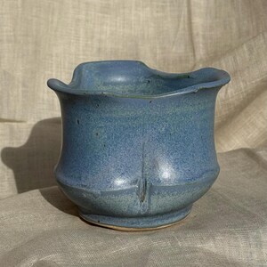 Op de afbeelding: Een handgemaakte, blauwe keramische vaas met een unieke, golvende rand. De vaas heeft een textuur en een licht uitlopende basis. De kleur is een mix van blauw en blauwgroen, met subtiele kleurschakeringen. Het is een decoratief item.