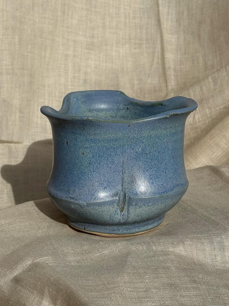 Op de afbeelding: Een handgemaakte, blauwe keramische vaas met een unieke, golvende rand. De vaas heeft een textuur en een licht uitlopende basis. De kleur is een mix van blauw en blauwgroen, met subtiele kleurschakeringen. Het is een decoratief item.