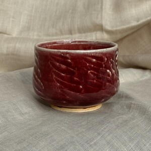 Op de afbeelding: Een handgemaakte, dieprode keramische kom met een getextureerde buitenkant. De kom heeft een brede, afgeronde vorm en een lichtgekleurde basis. De binnenkant is een lichtere tint rood. De kom wordt waarschijnlijk gebruikt om te serveren of als decoratief stuk.