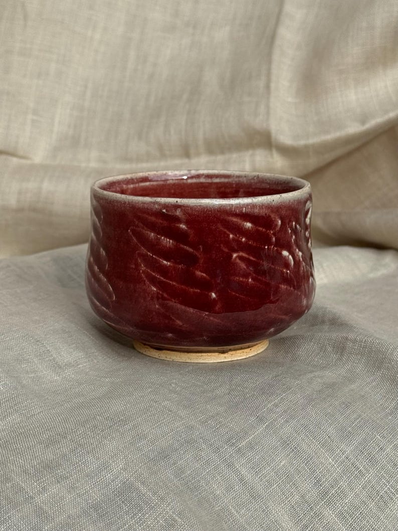 Op de afbeelding: Een handgemaakte, dieprode keramische kom met een getextureerde buitenkant. De kom heeft een brede, afgeronde vorm en een lichtgekleurde basis. De binnenkant is een lichtere tint rood. De kom wordt waarschijnlijk gebruikt om te serveren of als decoratief stuk.