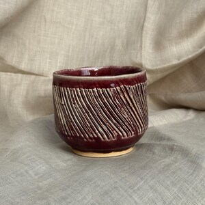 May include: A handmade ceramic cup with a deep burgundy glaze and textured diagonal lines. The cup has a rounded shape and a light beige base. The interior of the cup is also burgundy.