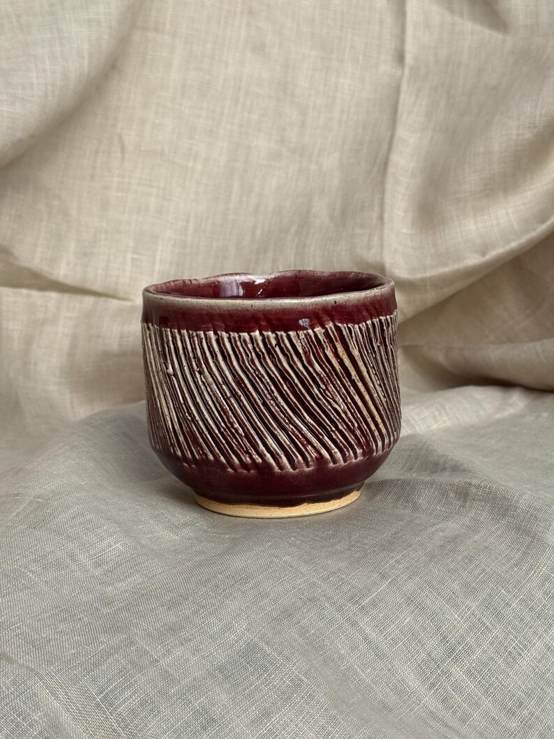Op de afbeelding: Een handgemaakte keramische beker met een diepe bordeauxrode glazuur en getextureerde diagonale lijnen. De beker heeft een afgeronde vorm en een lichtbeige basis. De binnenkant van de beker is ook bordeaux.