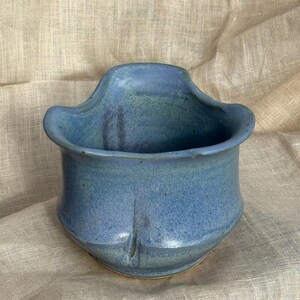 Op de afbeelding: Een handgemaakte, blauwe keramische plantenbak met een uniek, gebogen ontwerp. De plantenbak heeft een brede opening en een getextureerd oppervlak, perfect voor kleine planten. Het blauwe glazuur heeft subtiele variaties.