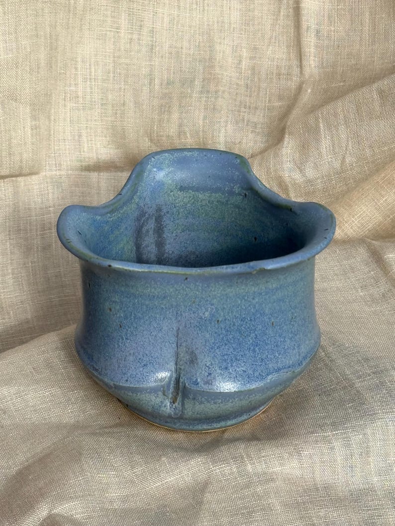 Op de afbeelding: Een handgemaakte, blauwe keramische plantenbak met een uniek, gebogen ontwerp. De plantenbak heeft een brede opening en een getextureerd oppervlak, perfect voor kleine planten. Het blauwe glazuur heeft subtiele variaties.