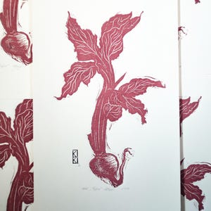 May include: A linocut print on white paper featuring a dark red botanical illustration of a root vegetable with leafy stems. The artwork includes a small black geometric signature mark on the left and handwritten text at the bottom centre.