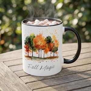May include: A white ceramic mug with a black rim and handle, featuring a watercolor illustration of autumn trees in shades of orange, yellow, and green. The text Fall Magic is printed in black script below the trees, and the mug contains hot cocoa.