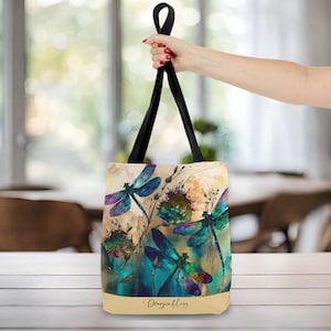 May include: A beige tote bag with black straps features a watercolor design of blue, green, and purple dragonflies on a beige background. The text "Dragonflies" is printed on the bottom of the bag.