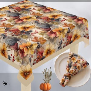May include: A square tablecloth featuring a watercolour-style print of autumn leaves, yellow flowers, and red berries on a light background. A matching folded napkin is placed on a white plate, accompanied by a small orange pumpkin and dried herbs.
