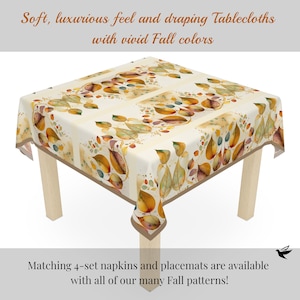 May include: A square tablecloth featuring a watercolour-style pattern of autumn leaves in shades of orange, yellow, and brown on a cream background. The text reads: Soft, luxurious feel and draping Tablecloths with vivid Fall colours. Matching 4-set napkins and placemats are available with all of our many Fall patterns!
