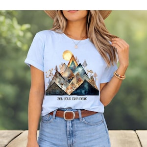 May include: A light blue short-sleeve t-shirt featuring a graphic print of stylized mountains with a golden sun and trees. The design includes the text "Set Your Own Peak" in black lettering centered below the mountain illustration.