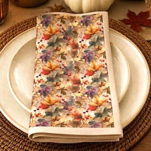 May include: A folded cloth napkin featuring a repeating pattern of autumn maple leaves in shades of orange, yellow, and red, interspersed with small red berries and muted green foliage, finished with a solid light beige border on all sides.