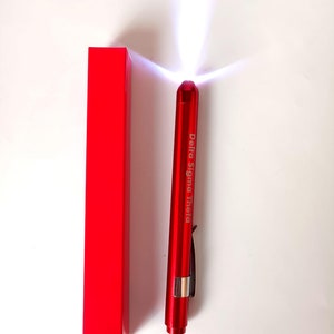 May include: A red penlight with the text "Delta Sigma Theta" printed on the side, next to a matching red rectangular box. The penlight is emitting a bright white light. The penlight has a silver clip.