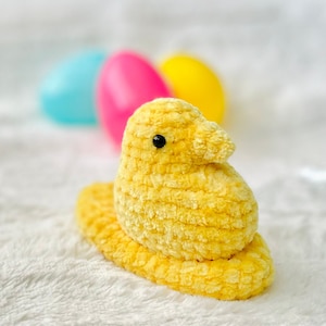 May include: A yellow plush chick with a black eye, sitting on a white surface. The chick is shaped like a traditional Easter candy.