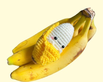 Könnte beinhalten: Eine gehäkelte Banane mit einem weißen Gesicht und schwarzen Augen, die auf einem Bund gelber Bananen liegt.