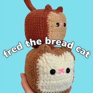 Può includere: Due peluche di gatto all'uncinetto impilati uno sopra l'altro. Il gatto in alto è marrone e beige, e il gatto in basso è beige e bianco. Il gatto in basso ha il testo "fred the bread cat" scritto davanti a lui.