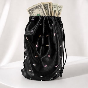 May include: Black satin drawstring bag filled with US dollar bills. The bag is adorned with scattered iridescent sequins. The drawstring is black, and the bag is sitting on a white surface.