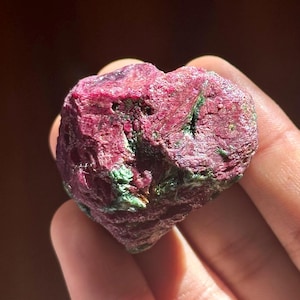May include: A close-up of a dark red ruby crystal with green inclusions. The crystal is rough and has a natural, unpolished appearance.