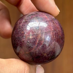 May include: A polished, spherical gemstone in shades of deep red and burgundy. The orb has a smooth, reflective surface, with visible internal patterns. The gemstone is held in a hand, showcasing its size and texture. This item is ideal for collectors or those interested in crystals.