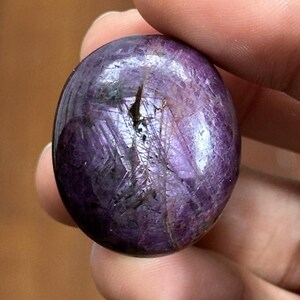 Puede incluir: Una piedra preciosa pulida, redonda y morada, con una superficie reflectante. La piedra tiene un aspecto oscuro y moteado con vetas más claras y una grieta. La gema se sostiene en una mano sobre un fondo de madera borroso.