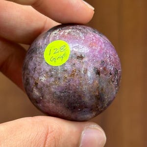 May include: A polished, spherical gemstone with a mottled appearance, featuring shades of purple, brown, and hints of pink. A small, round, yellow sticker with the text "128 G" is affixed to the surface. The gemstone is held in a hand.