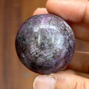 May include: A small, spherical gemstone with a mix of purple, gray, and black hues. The polished surface reflects light, showcasing a subtle, textured pattern. The gemstone is held in a hand, with a wooden background.