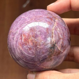 May include: A polished, spherical gemstone in shades of purple and pink, held in a hand. The orb has a swirling pattern and a glossy finish, reflecting light. This decorative sphere is a unique home decor item.