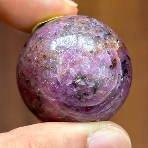 May include: A polished, spherical gemstone in shades of pink and purple, with dark speckles. The gemstone is held between two fingers, showcasing its smooth, rounded form. The gemstone is a decorative item.