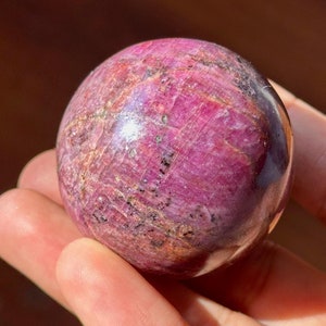 May include: A polished, spherical gemstone in shades of deep pink and burgundy, with subtle surface variations. The orb is held in a hand, showcasing its smooth, reflective surface. The gemstone's color and texture suggest a natural mineral specimen.