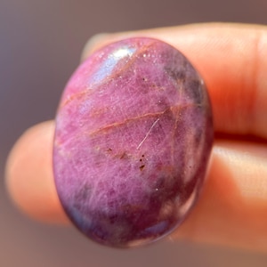 May include: A smooth, oval-shaped pink gemstone with a subtle shimmer. The stone has a deep pink hue with streaks of lighter pink and white.