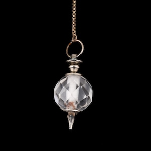 May include: A faceted crystal pendulum with a silver-colored metal cap and a pointed tip, suspended from a delicate gold-colored chain. The background is solid black.
