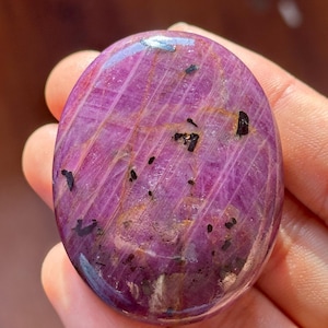May include: A smooth, oval-shaped purple gemstone with black and gold inclusions. The gemstone is polished and has a glossy finish.