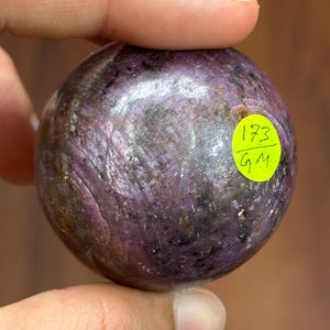 May include: A polished, spherical gemstone with a deep purple hue, featuring swirling patterns and a textured surface. A small, yellow sticker with the numbers "173" and the letters "GM" is affixed to the stone.