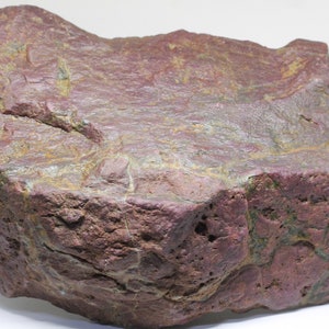 Puede incluir: Una roca grande, de forma irregular, con una superficie rugosa y texturizada. La roca muestra un color burdeos intenso con vetas amarillas y verdes. La apariencia general sugiere una piedra natural sin pulir, posiblemente un espécimen mineral.
