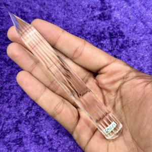 May include: A clear crystal point with a faceted design. The point is approximately 6.0 cm long and is held in a hand.