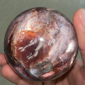 May include: A polished, spherical gemstone with a mix of red, brown, and white hues. The orb-shaped stone has a reflective surface, showcasing a natural, layered pattern. The gemstone is held in a hand, highlighting its size and texture.