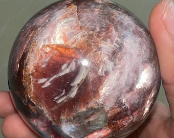 May include: A polished, spherical gemstone with a mix of red, brown, and white hues. The orb-shaped stone has a reflective surface, showcasing a natural, layered pattern. The gemstone is held in a hand, highlighting its size and texture.