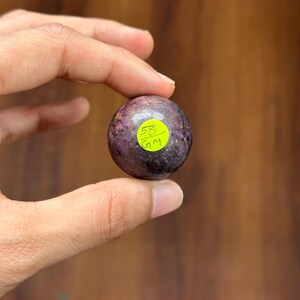 May include: A small, round, polished stone with a mottled purple and gray appearance. A yellow sticker with the number "58" and the letters "GM" is affixed to the stone. The stone is held between fingers.