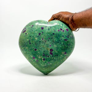 Puede incluir: Una gran escultura de piedra en forma de corazón. La piedra es de color verde moteado con inclusiones de color púrpura intenso. La escultura está sostenida por una mano y el fondo es blanco.