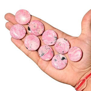 Puede incluir: Una mano sosteniendo una colección de piedras rosas y redondas. Las piedras tienen un aspecto veteado con inclusiones grises y negras más oscuras. Las piedras son lisas y pulidas, con una estética natural y terrosa.